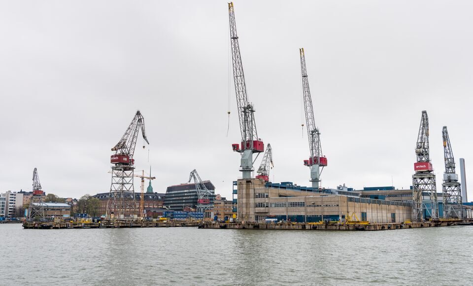 An industrial harbor shipyard scene featuring towering cranes, buildings and stacks of containers.