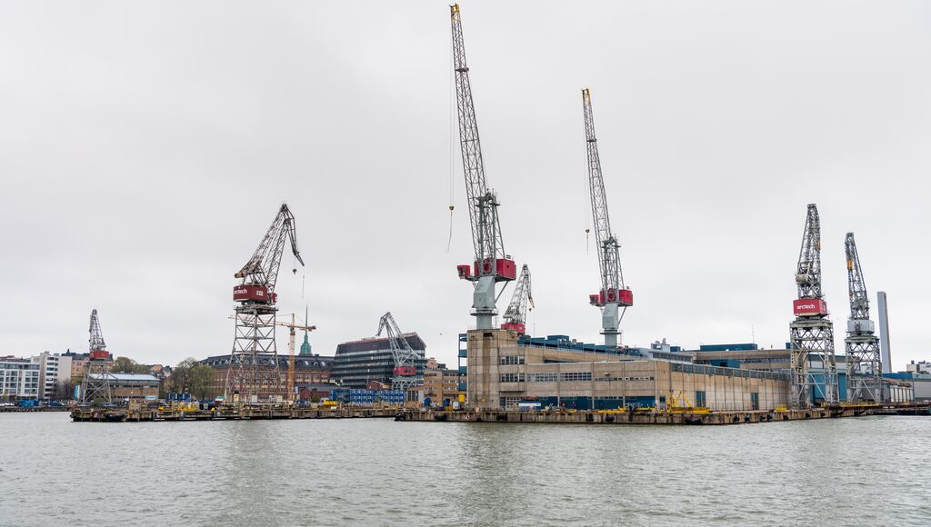 An industrial harbor shipyard scene featuring towering cranes, buildings and stacks of containers.