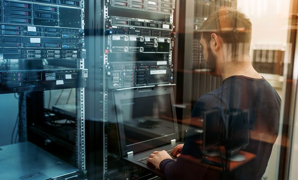 A man typing on a laptop in a data center environment.