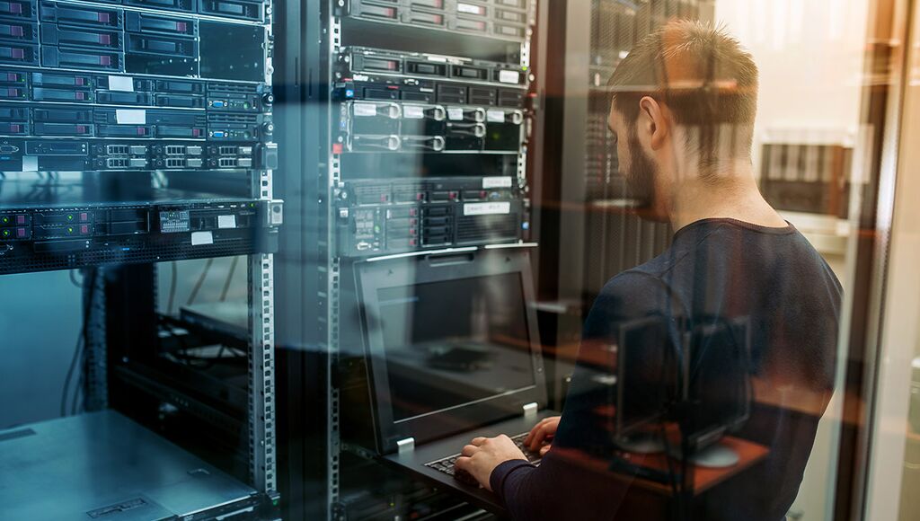 A man typing on a laptop in a data center environment.